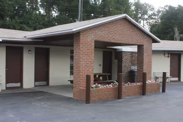 Exterior view of a single-story building with beige walls and brown doors, featuring a covered brick entrance area with a picnic table and flower bed. The building is surrounded by trees and has a paved driveway in front.