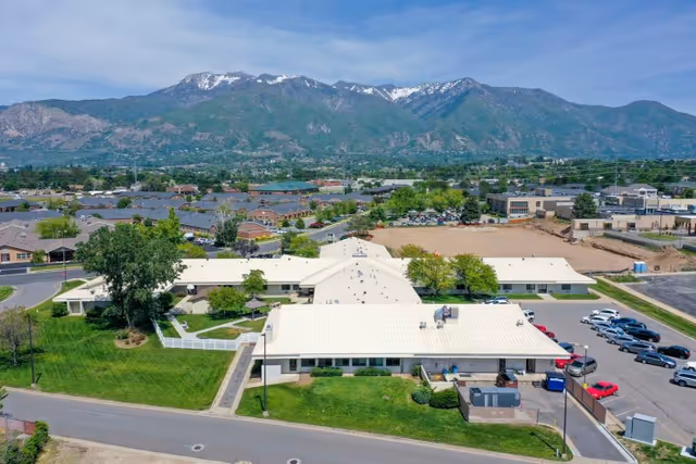 Aerial view of Mt. Ogden Health And Rehabilitation Center, a single-story building with a white roof surrounded by green lawns, parking lots, and trees. In the background, there are residential neighborhoods and a mountain range with some snow on the peaks under a partly cloudy sky.