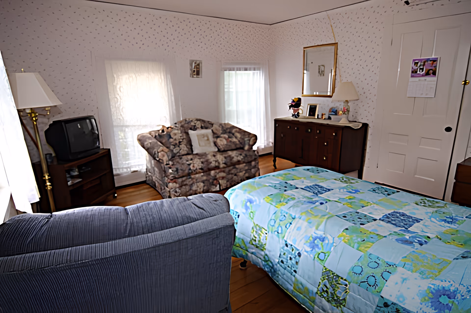 A cozy bedroom with a bed covered in a colorful patchwork quilt, a floral loveseat, a blue armchair, a wooden dresser with a mirror, a floor lamp, and an old-style television on a stand. The room has two windows with sheer white curtains and wallpaper with a subtle pattern.
