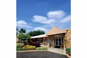 Exterior view of the Ross Center for Nursing and Rehabilitation building with a clear blue sky and some clouds. The entrance has a covered walkway with a triangular roof, surrounded by landscaped bushes and greenery.