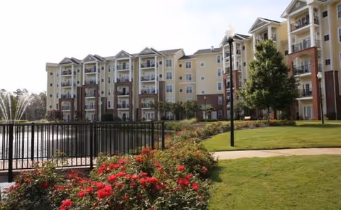 Multi-story senior living building with balconies, landscaped grounds, and a paved walkway.