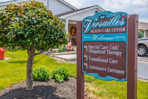 Exterior view of Versailles Health Care Center entrance with a decorative sign and trimmed landscaping.