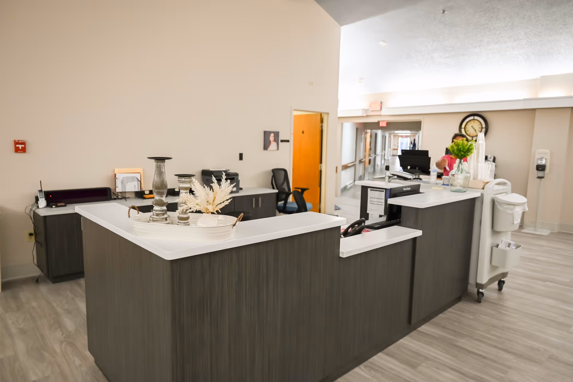 Reception desk area inside a senior care facility with a modern dark wood counter, decorative items on the counter, office chair, computer monitors, and a hallway extending in the background.