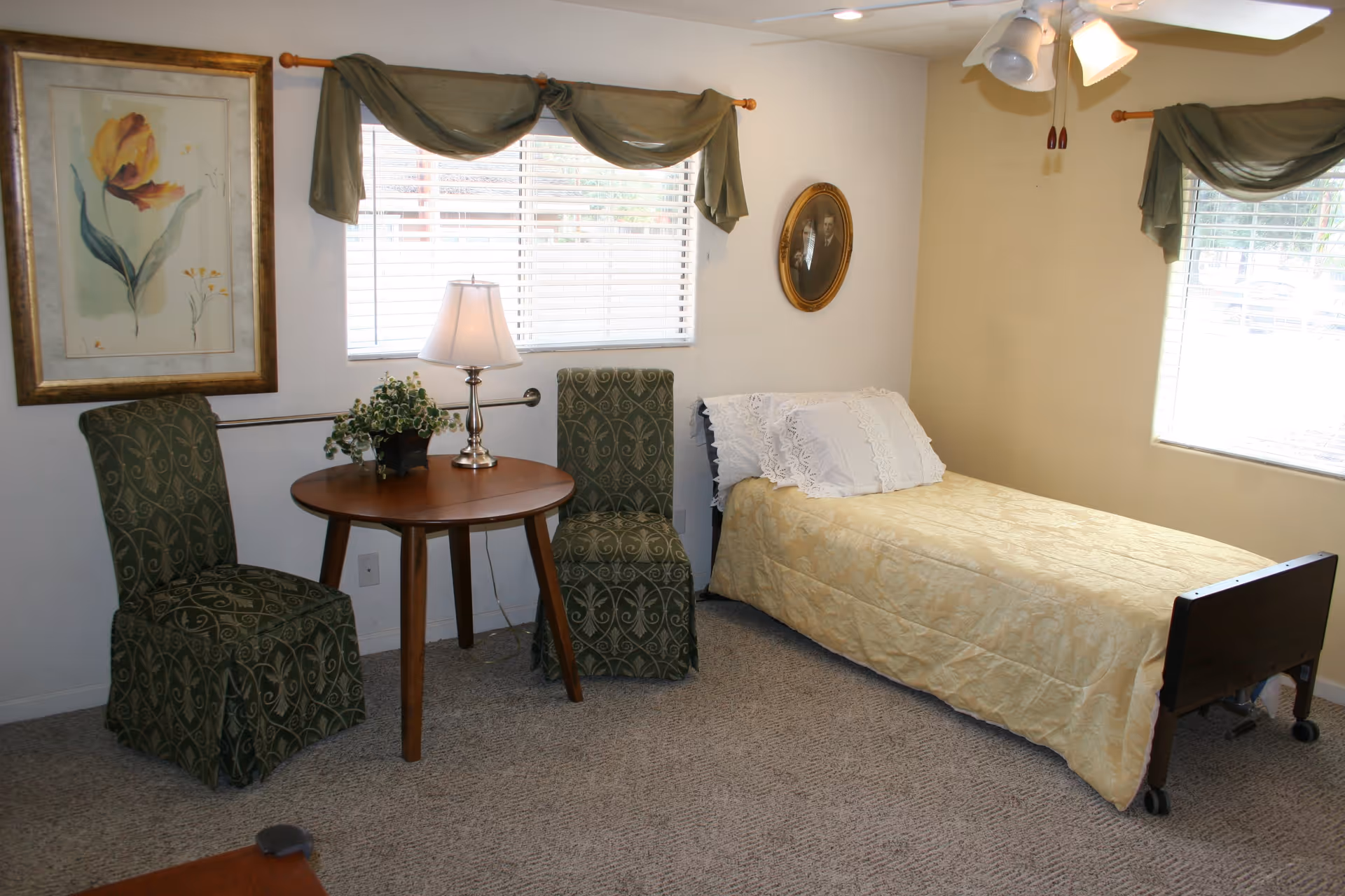 A small, cozy bedroom with a single bed covered in a yellow patterned bedspread and white lace pillows. Next to the bed are two green patterned chairs and a round wooden table with a lamp and a small plant. The room has two windows with green draped curtains, a ceiling fan with lights, a framed floral painting on one wall, and an oval framed black-and-white photograph on another wall.