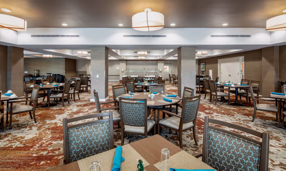 A spacious dining room with multiple tables and chairs arranged neatly. Each table is set with placemats, glasses, and turquoise napkins. The room has a patterned carpet in shades of brown and beige, modern ceiling lights, and neutral-colored walls. There are columns in the center and a buffet area at the back.
