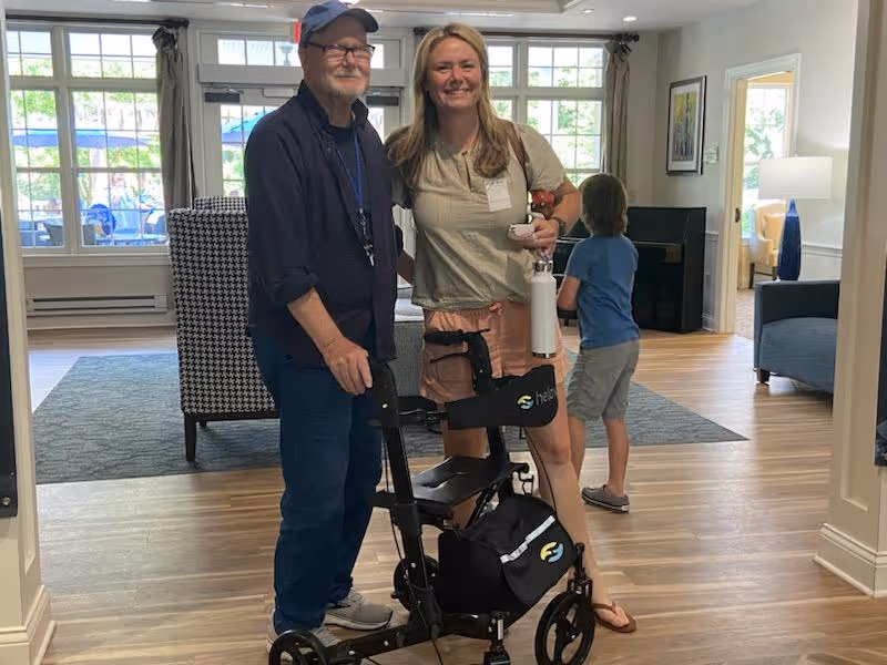 An elderly man and a woman standing together inside a well-lit living area with wooden floors. The man is using a black walker with a seat and storage bag, and both are smiling at the camera. In the background, a young boy is walking away, and there is furniture including chairs, a lamp, and framed artwork on the walls.