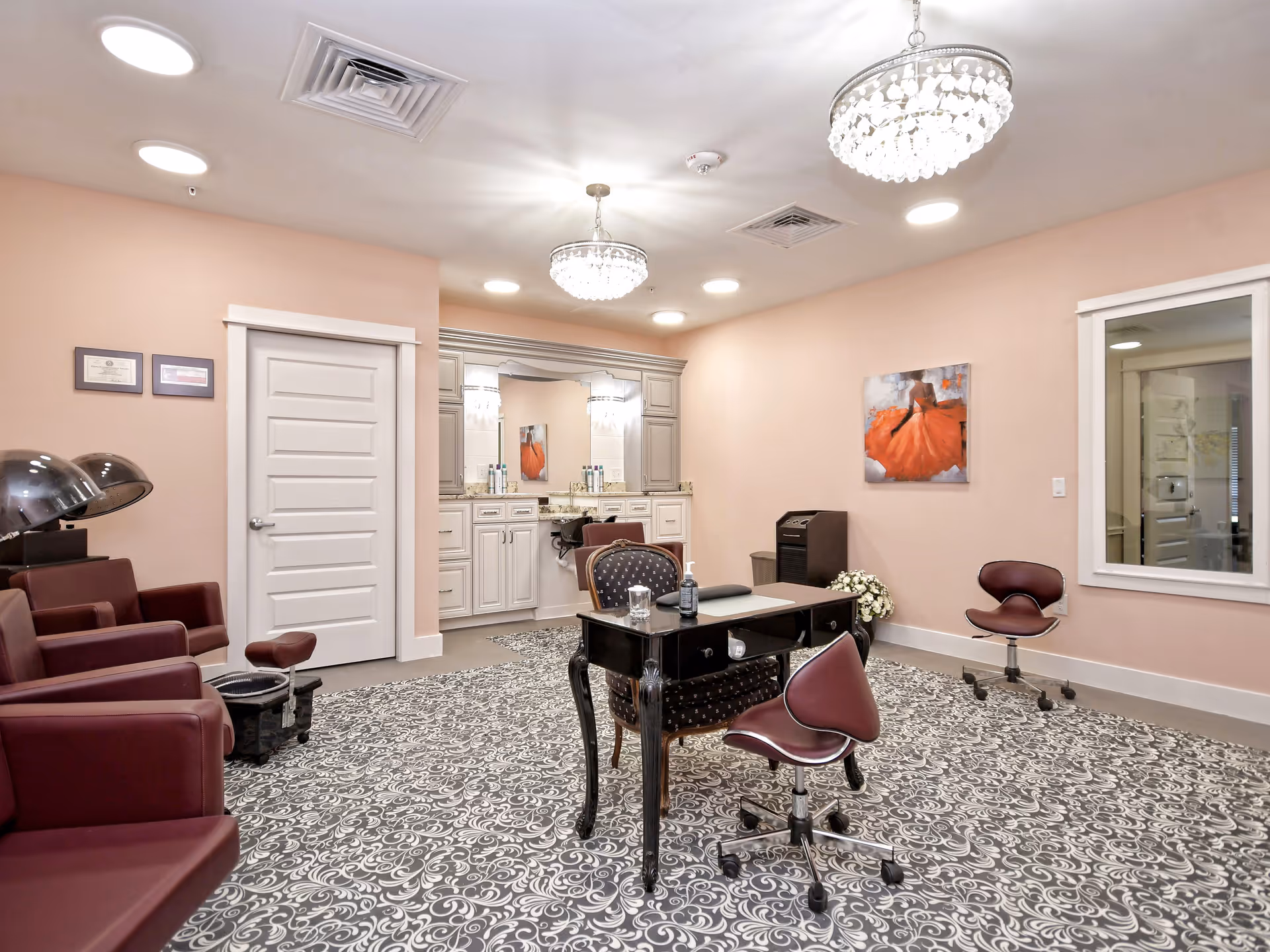 A well-lit salon room with soft pink walls and patterned carpet. The room features three maroon salon chairs with hair dryers on the left, a black manicure table with two maroon rolling chairs in the center, and a large mirror with cabinetry and a sink at the back. Two crystal chandeliers hang from the ceiling, and a painting of a woman in an orange dress is on the right wall.