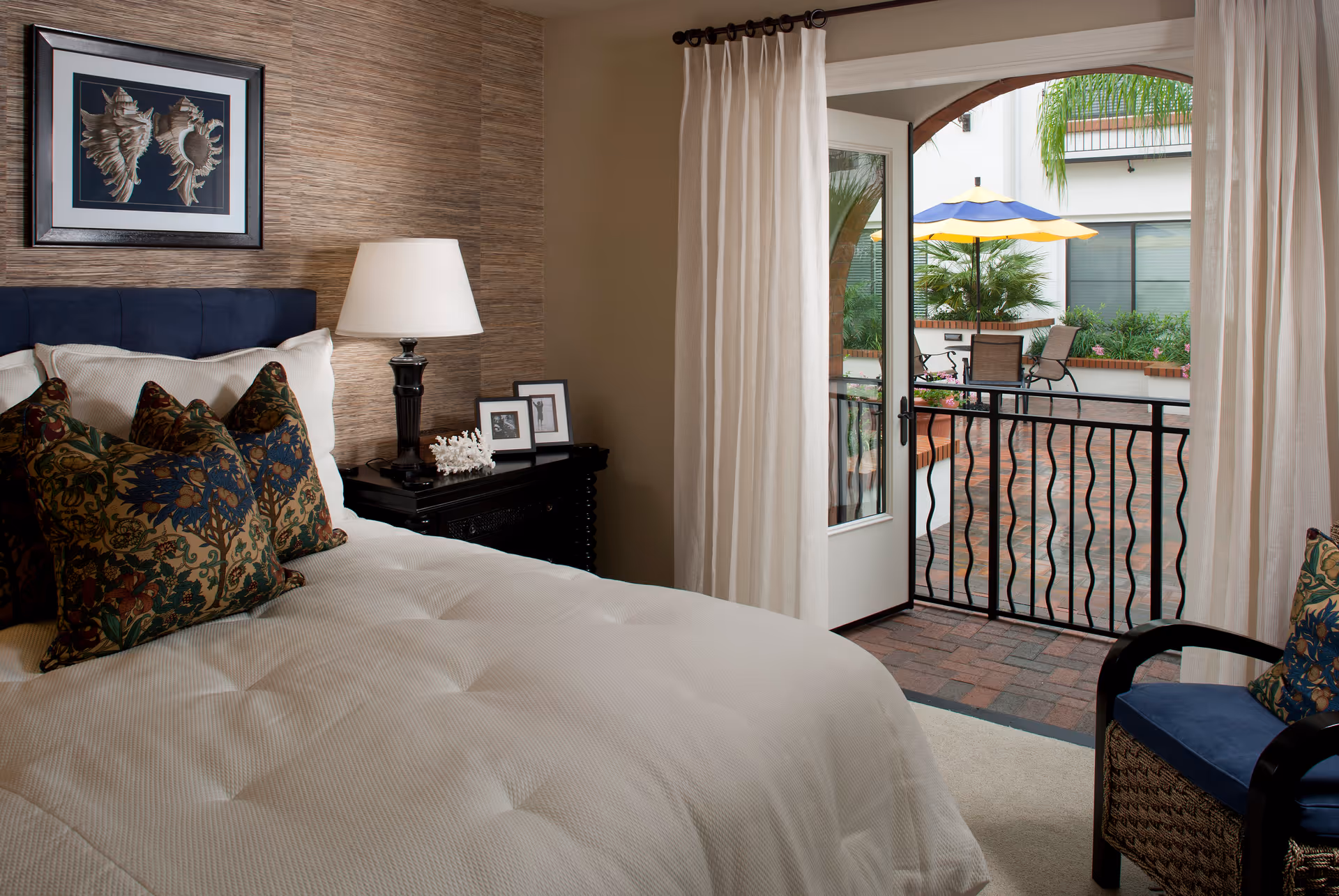 A cozy bedroom with a neatly made bed featuring decorative pillows, a bedside table with a lamp and framed photos, and an open door leading to a patio area with outdoor seating and a yellow and blue umbrella.