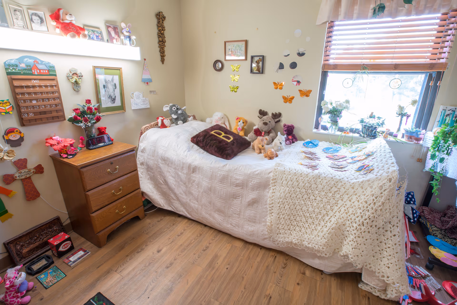 Small decorated bedroom with a single bed covered in a white quilt and crocheted throw, stuffed animals on the bed, a wooden nightstand, wall decorations, and a window with plants.