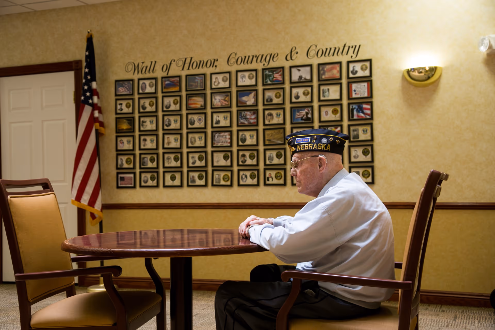 An elderly man wearing a Nebraska veteran cap sits alone at a round wooden table in a room with beige walls. Behind him is a wall display titled 'Wall of Honor, Courage & Country' featuring numerous framed photos and medals. An American flag stands in the corner near a closed door.