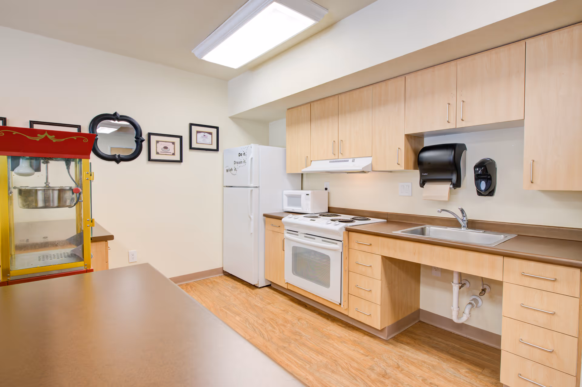 A clean and well-lit kitchen area with light wood cabinets, a white refrigerator, a white stove with oven, a microwave, a stainless steel sink, and a red and yellow popcorn machine. The walls are decorated with a few framed pictures and a black-framed mirror.