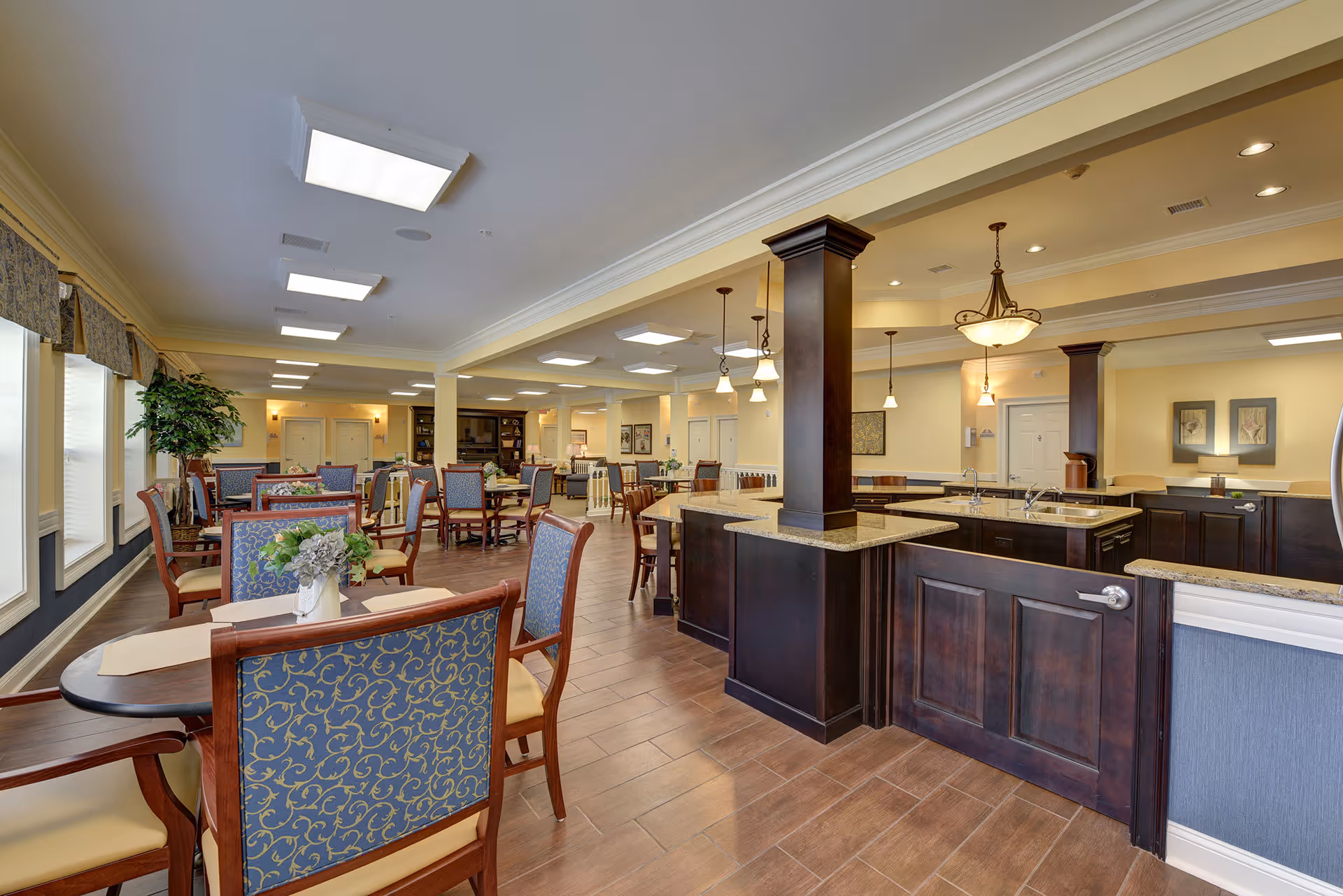 Spacious dining area in a senior living facility with multiple tables and chairs arranged neatly. The room features large windows with valances, wooden flooring, and a kitchen or serving counter with dark wood cabinetry and granite countertops. Pendant and ceiling lights illuminate the space, and there are decorative plants and framed artwork on the walls.