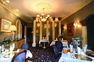 Elegant dining room with multiple tables covered in white tablecloths, set with napkins and glassware. The room features ornate columns, a chandelier, wall sconces, floral arrangements, and classic framed artwork on the walls. The carpet has a dark floral pattern, and the lighting creates a warm ambiance.