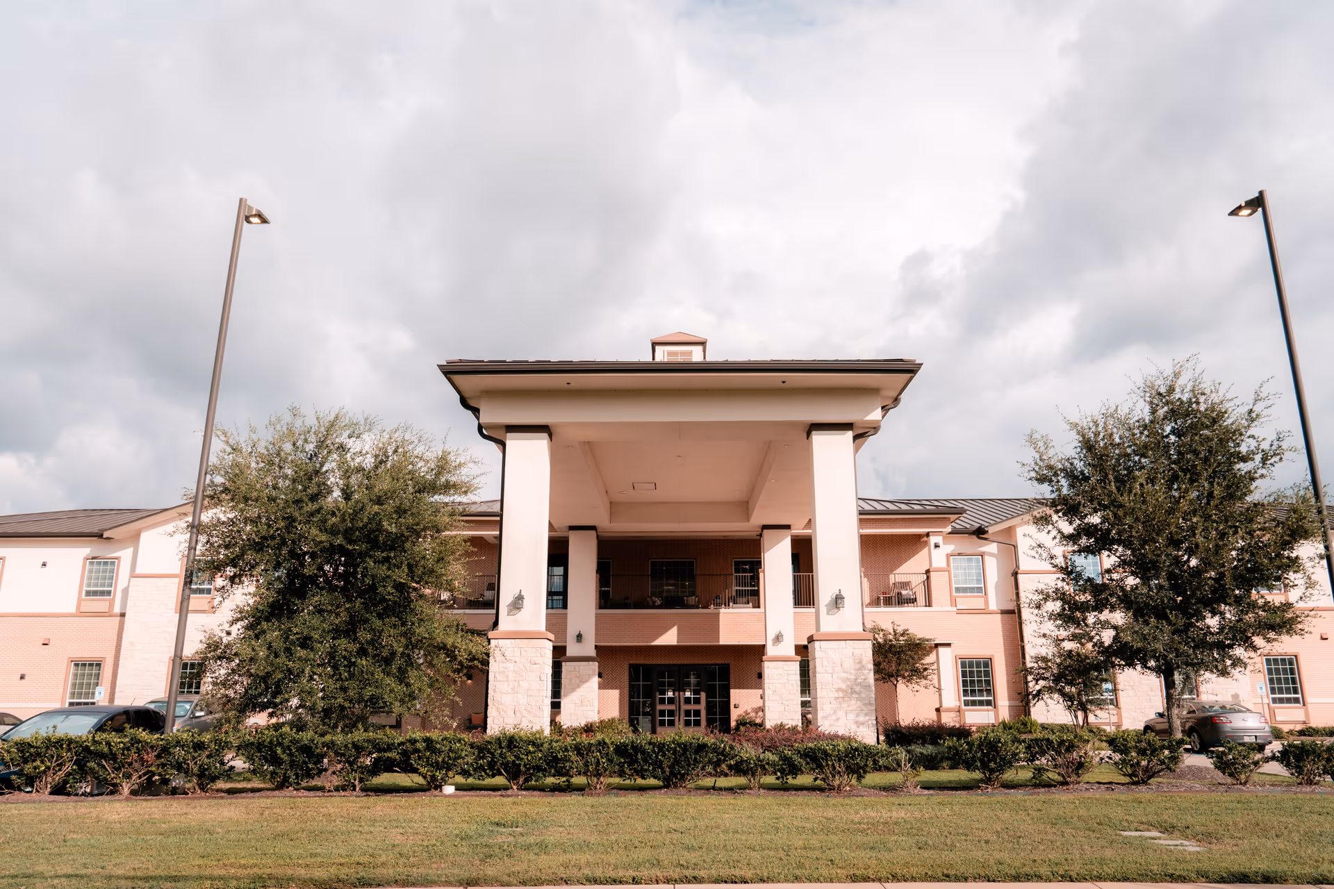 Front exterior view of a two-story senior living facility building with a covered entrance supported by large columns, surrounded by trees, shrubs, and a grassy lawn under a cloudy sky.
