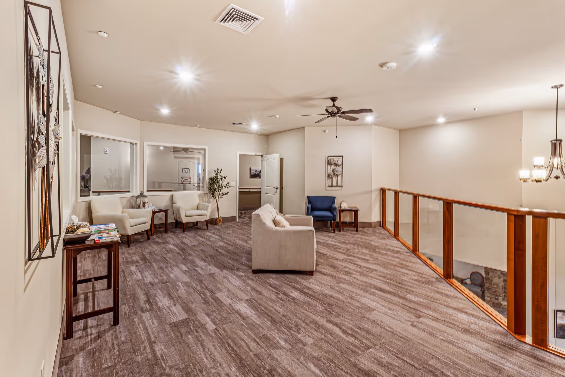 A spacious, well-lit common area with wood-style flooring and beige walls. The room features several armchairs in cream and blue colors arranged near small wooden tables. There is a ceiling fan with lights, a chandelier, and a wooden railing with glass panels overlooking a lower level. Decorative artwork and a potted plant add to the cozy atmosphere.