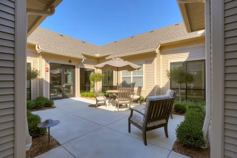 Outdoor patio area at American House Freedom Place Rochester with cushioned chairs, a round table, and an umbrella surrounded by beige building walls and windows under a clear blue sky.