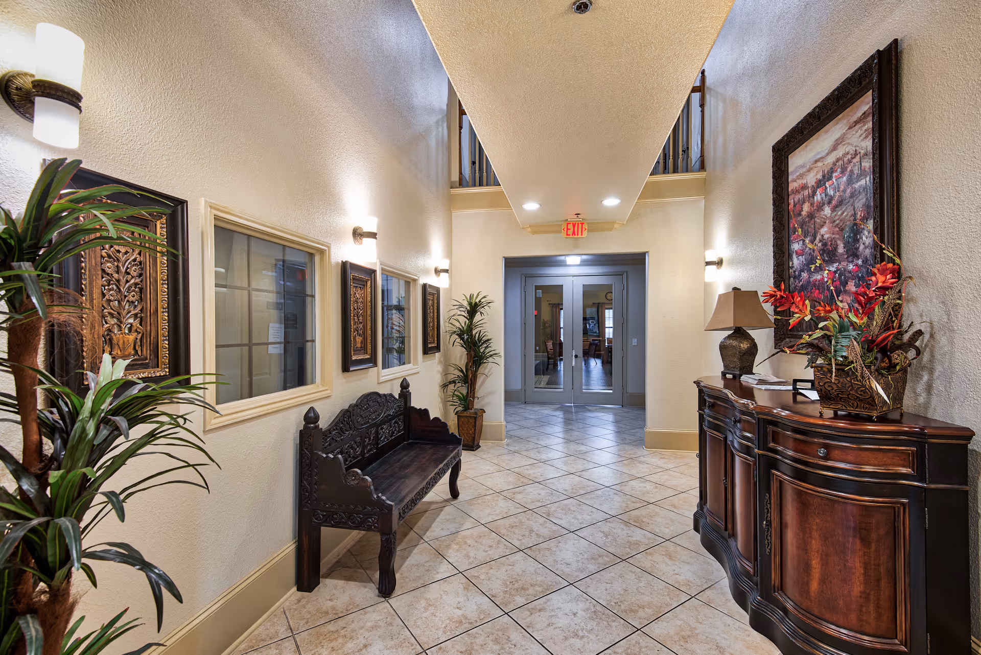 Interior hallway of a senior living facility with tiled floor, cream-colored walls, decorative wooden bench, potted plants, framed artwork, and a wooden sideboard with a lamp and floral arrangement. Double glass doors are visible at the end of the hallway.