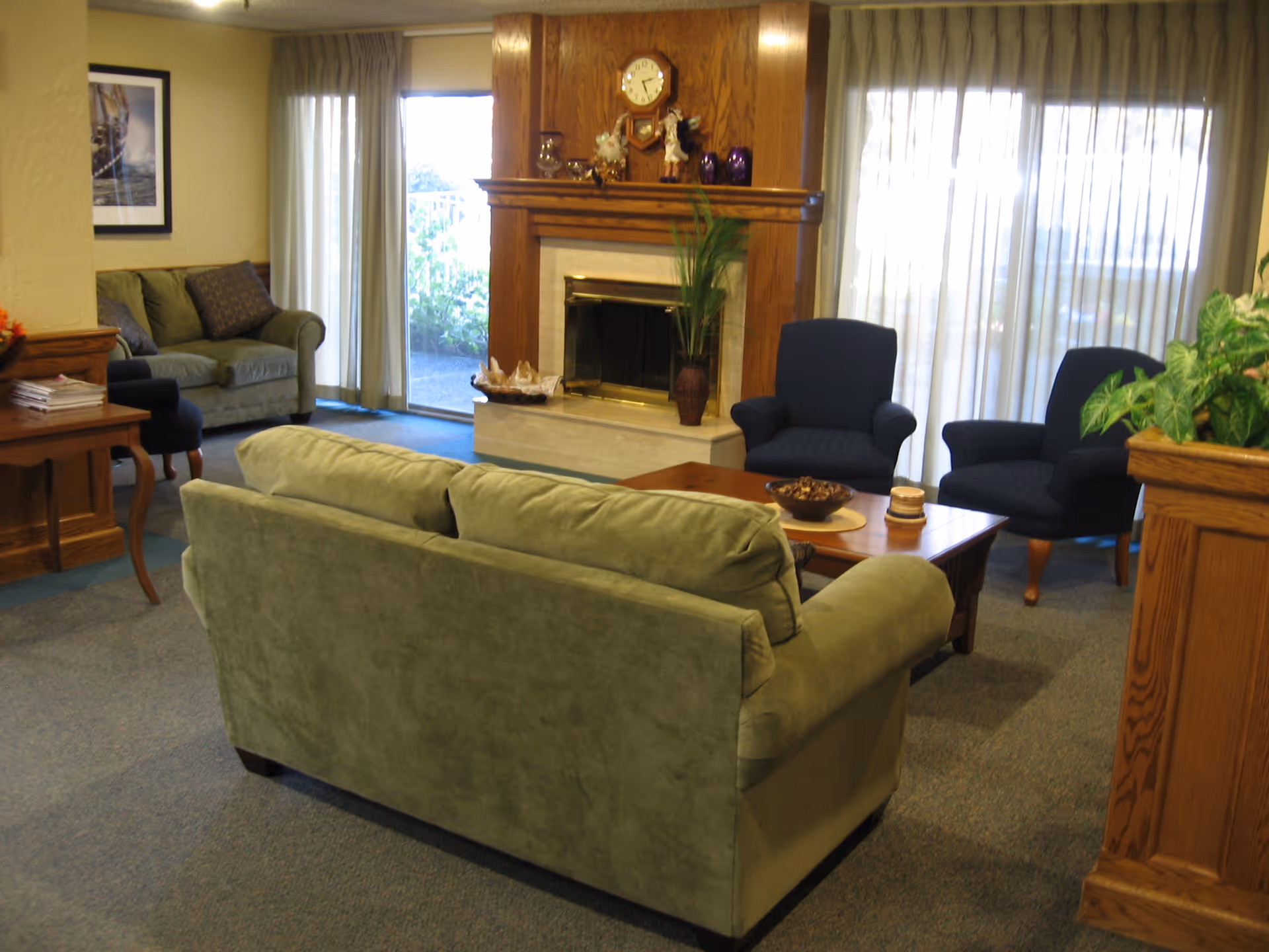 A cozy living room area with a green sofa facing a wooden coffee table and two dark blue armchairs. Behind the seating area is a fireplace with a wooden mantel decorated with a clock, figurines, and vases. Large windows with sheer curtains allow natural light to fill the room. There is also a side table with magazines and a framed picture on the wall.