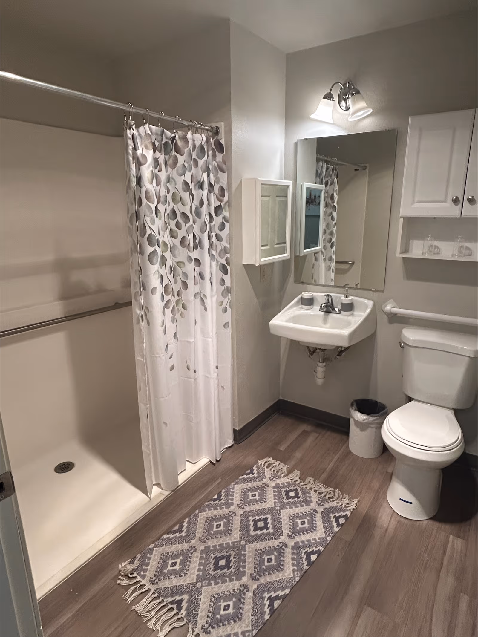 A clean and simple bathroom with a walk-in shower featuring a white curtain with a leaf pattern. The bathroom has a white sink with a mirror above it, a white toilet, a small white cabinet with glassware, a trash bin, and a patterned rug on the wooden floor.
