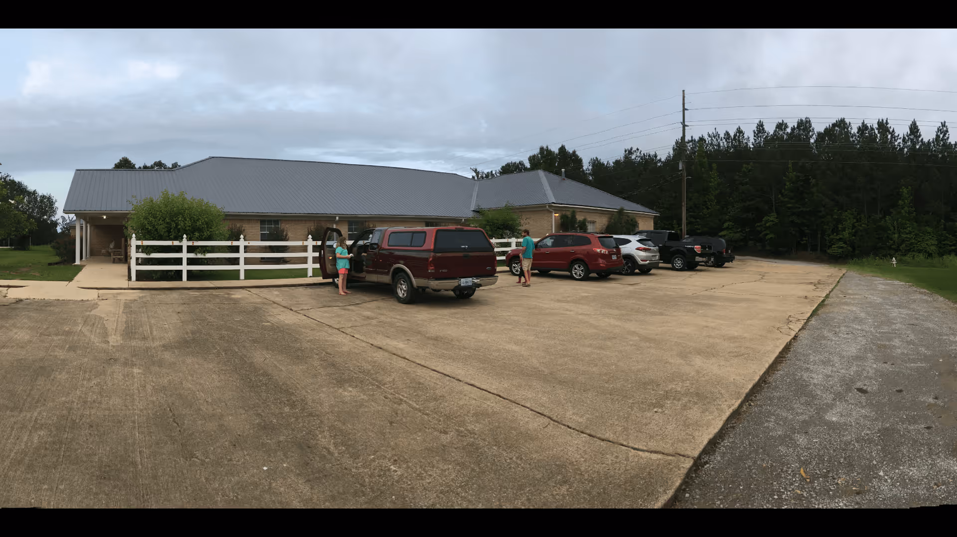 Exterior view of a single-story brick building with a gray metal roof, a white fence, and a concrete parking lot with several parked vehicles. Two people are standing near a red truck in the parking lot. Trees and power lines are visible in the background under a cloudy sky.
