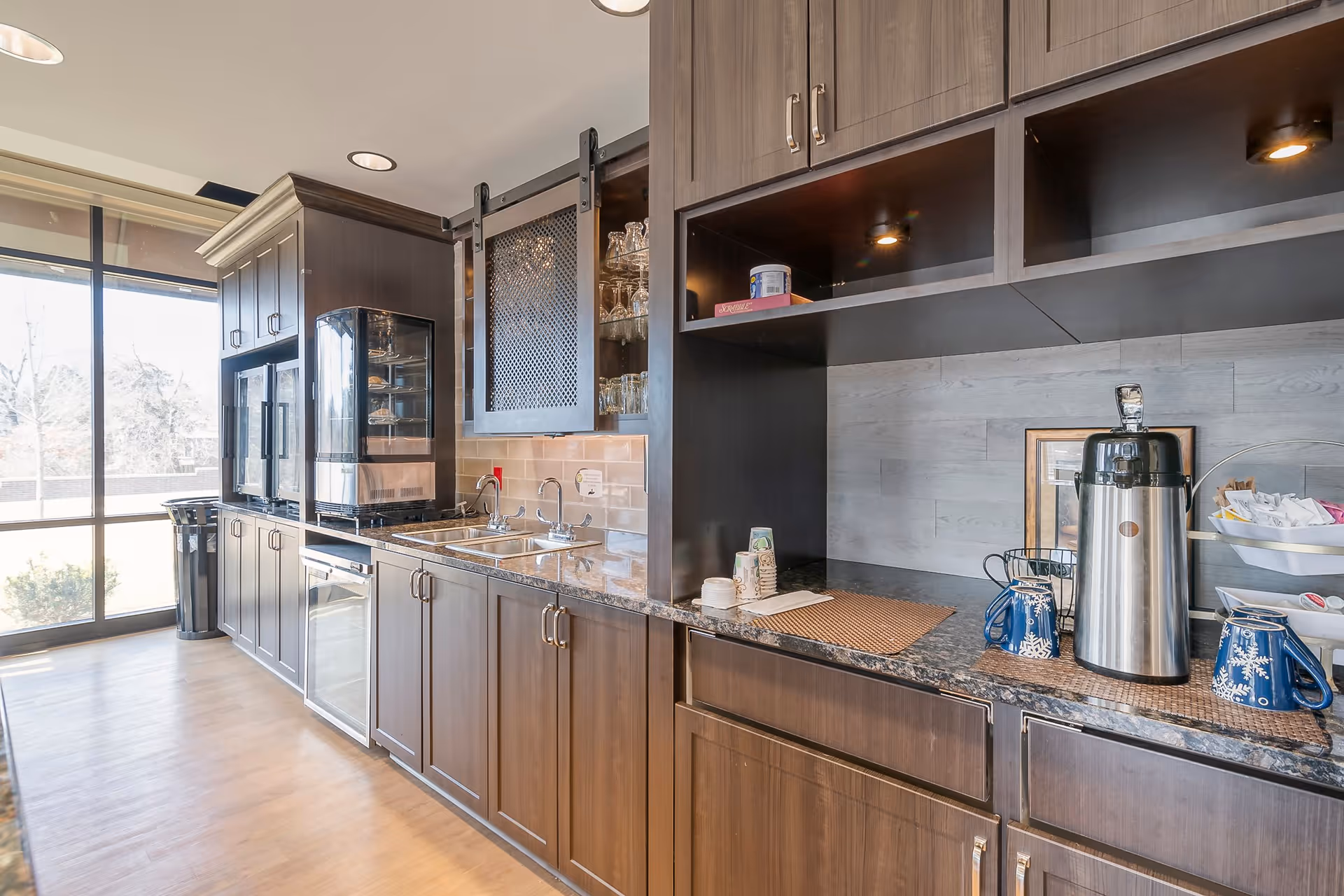 A modern kitchen area with dark wood cabinets, a granite countertop, a double sink, a coffee dispenser, and various cups and condiments. Large windows on the left side allow natural light to fill the space.