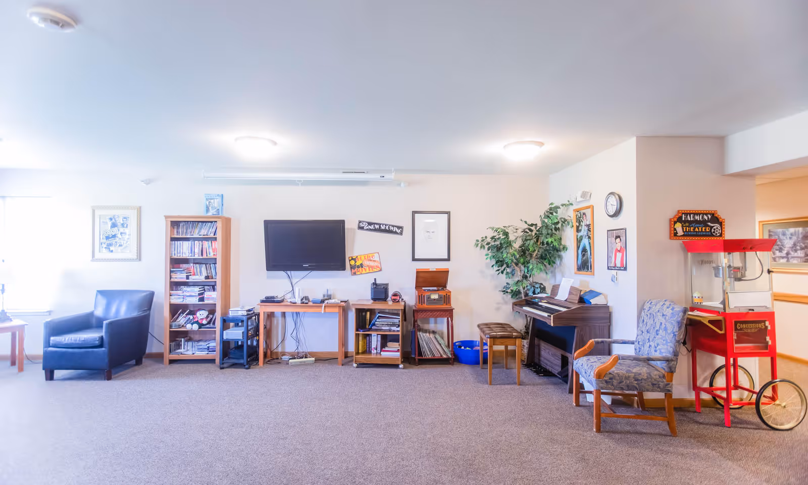 A bright and spacious common area in an assisted living facility featuring a black armchair, a wooden bookshelf filled with books and DVDs, a wall-mounted flat-screen TV, a wooden table with electronic devices, a vintage record player, a piano with a bench, a leafy green plant, framed pictures on the walls, a blue patterned armchair, and a red popcorn machine labeled 'Concessions'.