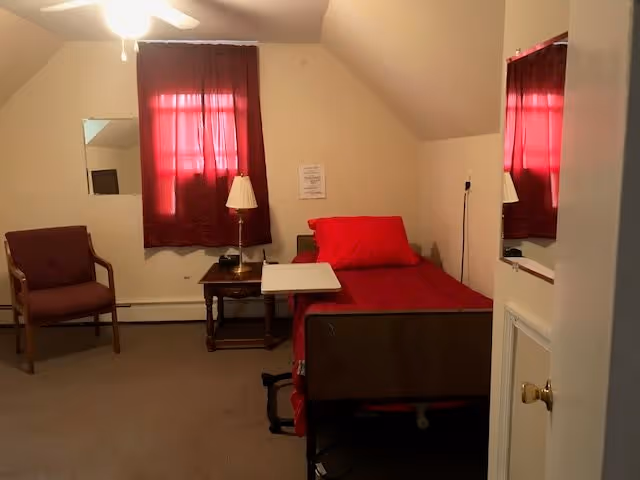 A small bedroom with a single bed covered in red bedding, a wooden nightstand with a lamp, a chair with armrests, a window with red curtains, and a ceiling fan. There is a mirror on the wall and a door partially open on the right side.