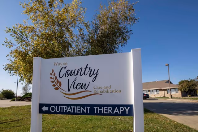 A freestanding white sign for 'Wayne Country View Care and Rehabilitation' with an 'OUTPATIENT THERAPY' arrow on a lawn in front of a building under a clear blue sky.