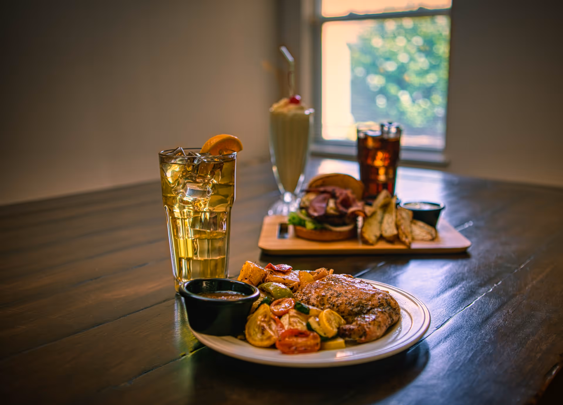 A wooden table with a plate of grilled chicken, roasted vegetables, and a small bowl of sauce in the foreground. Behind it, there is a glass of iced tea with a lemon wedge, a sandwich with potato wedges on a wooden board, a glass of cola, and a milkshake with whipped cream and a cherry. A window with blurred greenery outside is in the background.