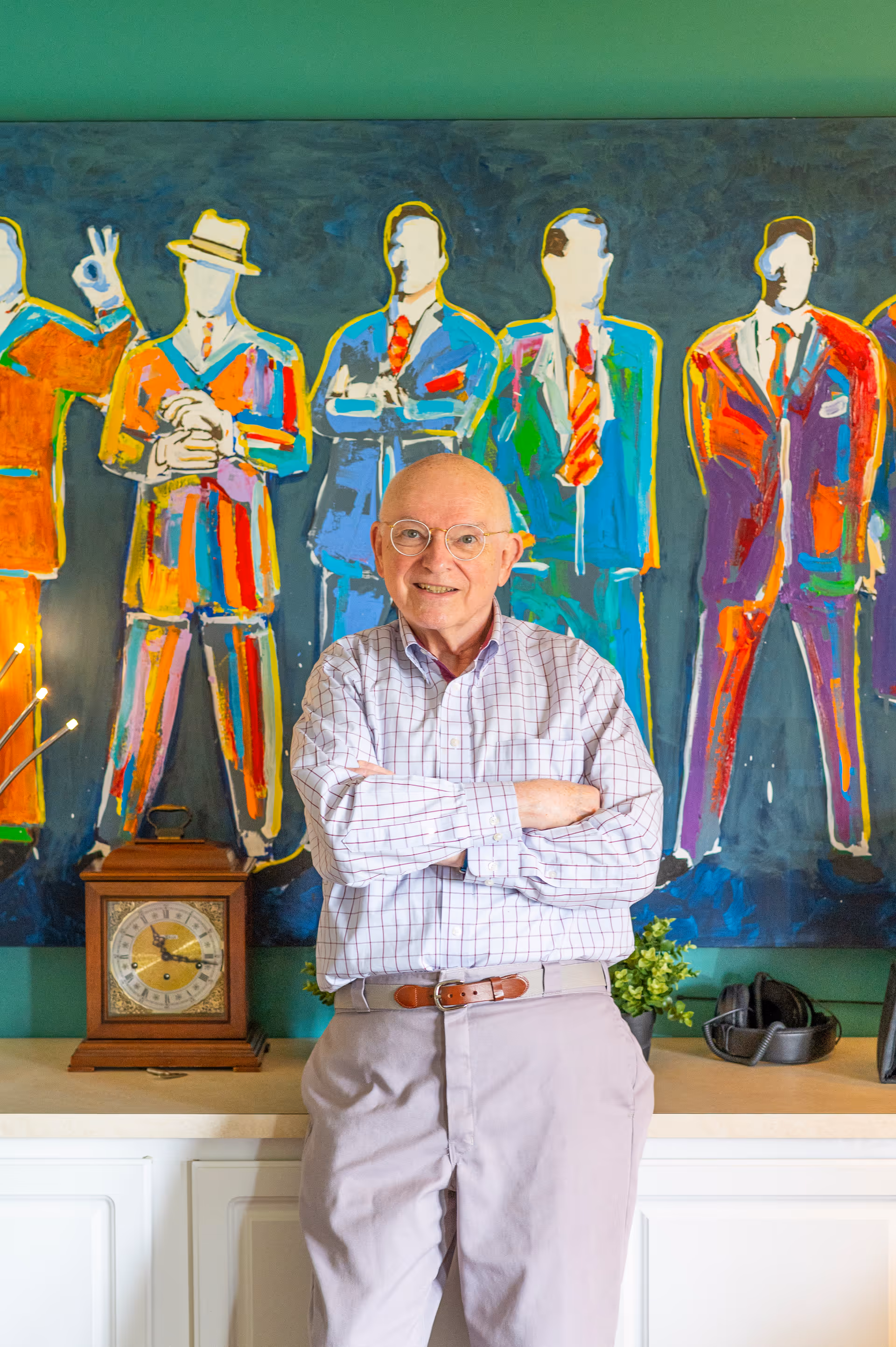 An elderly man with glasses and a checkered shirt stands with arms crossed in front of a colorful painting of faceless men in suits and hats. Behind him is a wooden clock and a small plant on a white cabinet against a green wall.