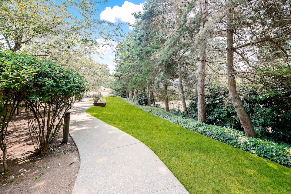 A paved walkway curves through a landscaped outdoor area with green grass, bushes, and tall trees under a partly cloudy blue sky.