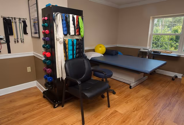 A therapy or exercise room with a padded treatment table, a black chair, and a rack holding colorful dumbbells and various exercise straps. The room has wooden flooring, beige and brown walls, and a window showing greenery outside.