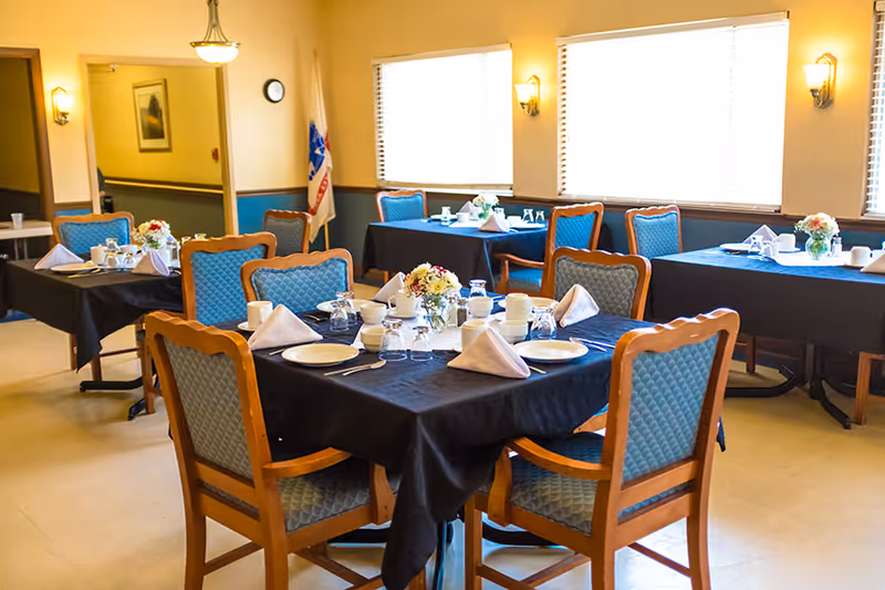 A dining room with multiple tables set with black tablecloths, place settings, folded napkins and floral centerpieces surrounded by upholstered chairs.