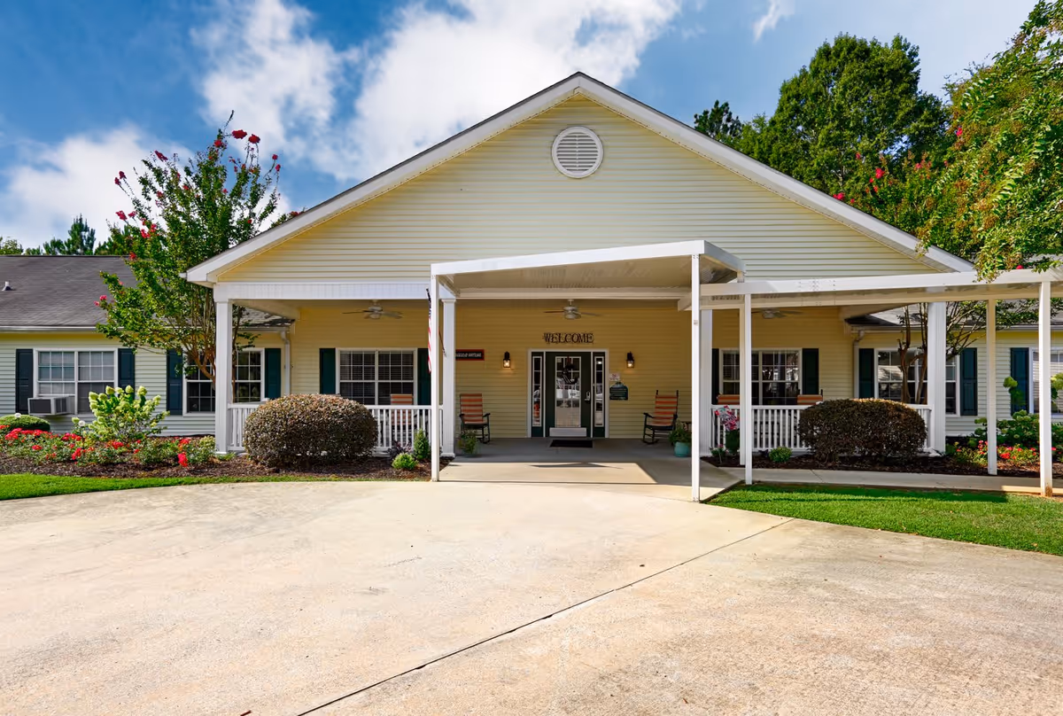 Front exterior view of a single-story yellow building with a covered entrance, rocking chairs on the porch, landscaped bushes and flowers, and a clear blue sky with some clouds.