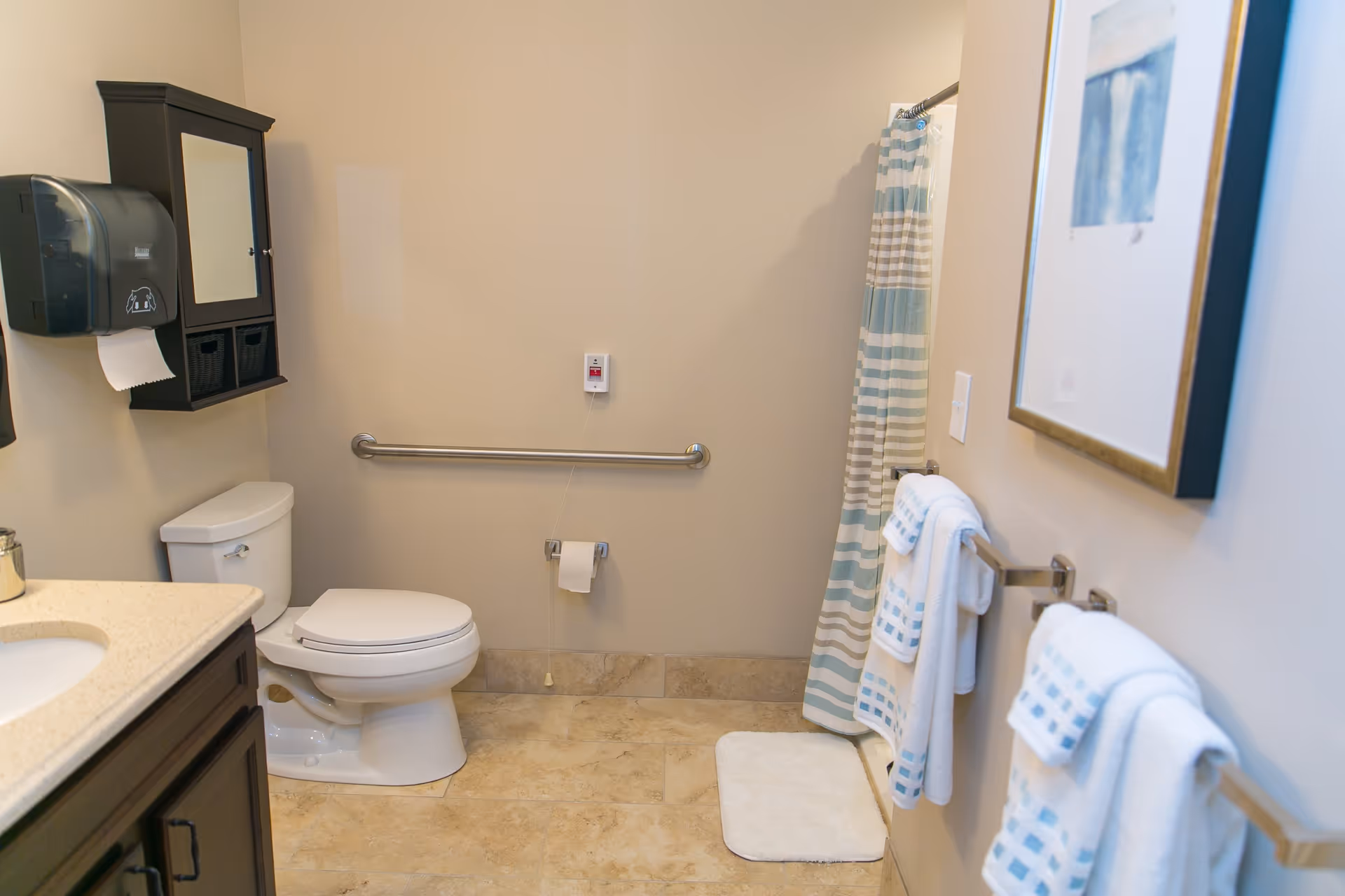 A clean and accessible bathroom featuring a white toilet with a grab bar on the wall behind it, a toilet paper holder, a shower with a striped curtain, a vanity with a sink and countertop, a wall-mounted cabinet with a mirror, a paper towel dispenser, and several white towels hanging on towel racks. The floor is tiled and there is a small white bath mat in front of the shower.