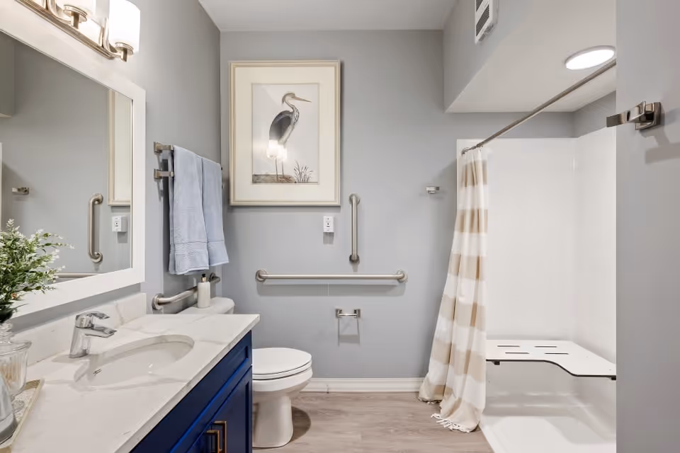 Accessible bathroom with a vanity sink, toilet, wall grab bars, and a shower with a bench and striped curtain.