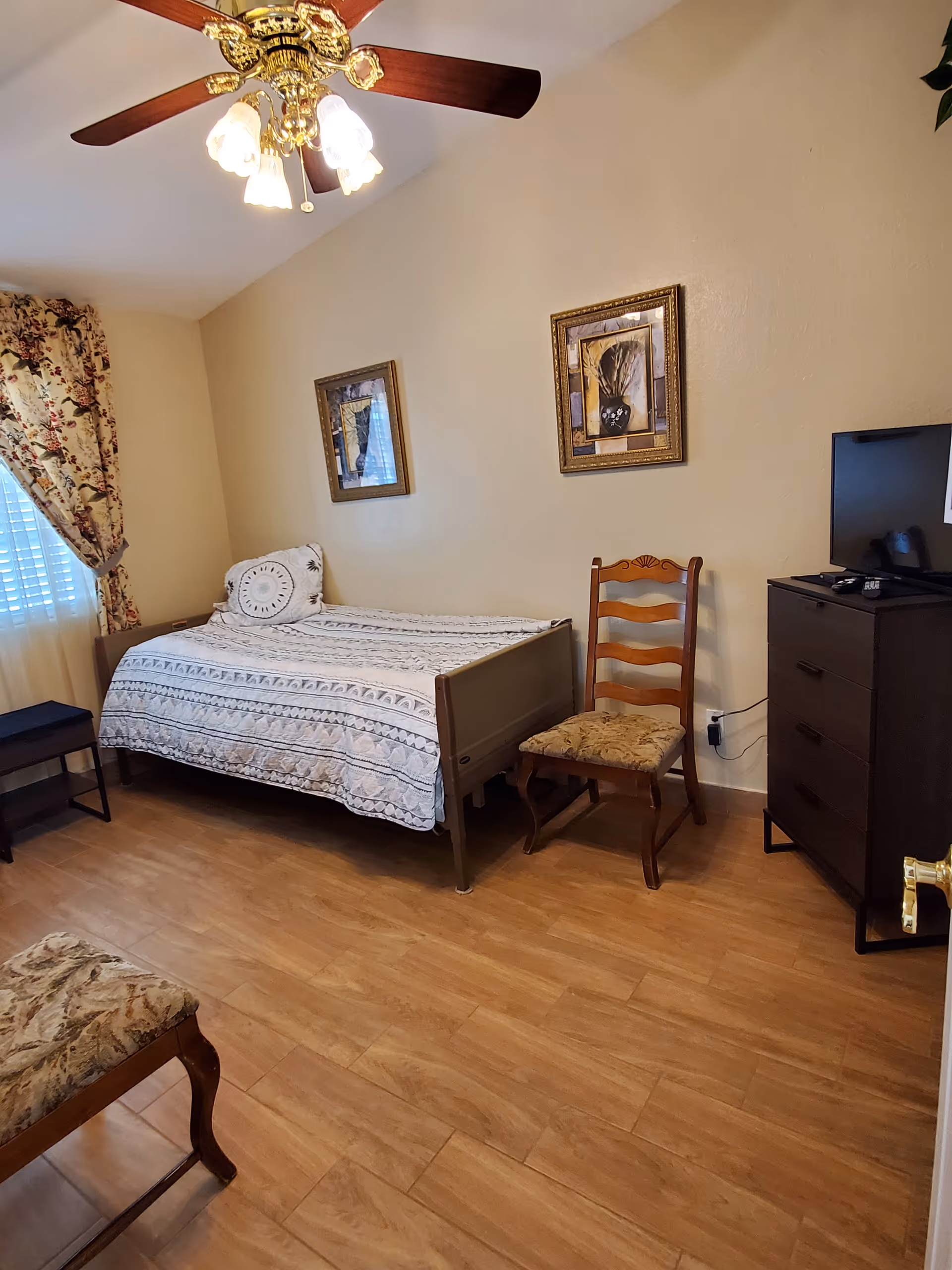 A bedroom with a single bed covered with a patterned bedspread, a wooden chair with a cushioned seat, a dark wooden dresser with a TV on top, two framed pictures on the wall, floral curtains on the window, and a ceiling fan with lights.