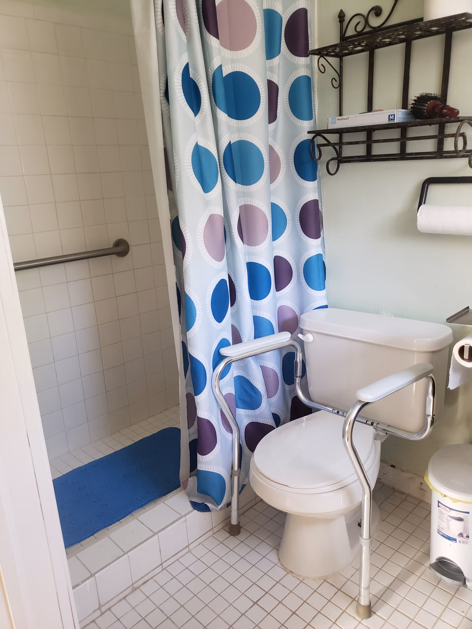 A bathroom with a white toilet equipped with arm supports for assistance, a shower area with a blue and purple polka dot shower curtain, a blue bath mat, a wall-mounted shelf holding a box and a hairbrush, a paper towel holder, and a small trash can on the tiled floor.