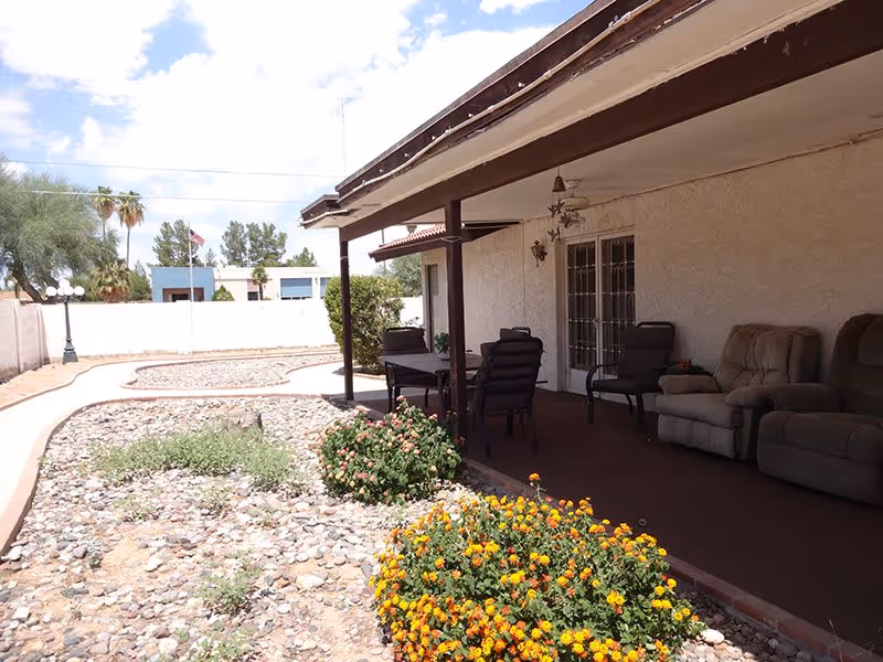 Outdoor patio area at The Sanctuary of Scottsdale with cushioned chairs and a table under a covered porch. The patio overlooks a garden with flowering plants and a rock bed, with a white wall and trees in the background under a partly cloudy sky.
