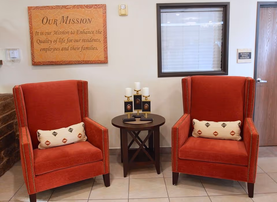 Two red upholstered armchairs with decorative pillows are placed on either side of a small round wooden table holding three candle holders with white candles. Behind the chairs, there is a window with closed blinds and a wooden door with a sign that reads 'Business Office Manager'. A framed sign on the wall reads 'Our Mission: It is our Mission to Enhance the Quality of life for our residents, employees and their families.'