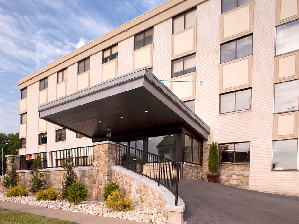 Exterior view of a multi-story brick building with a covered entrance featuring stone pillars and a ramp with black metal railings. There are small shrubs and landscaping with white rocks along the front of the building under a partly cloudy sky.