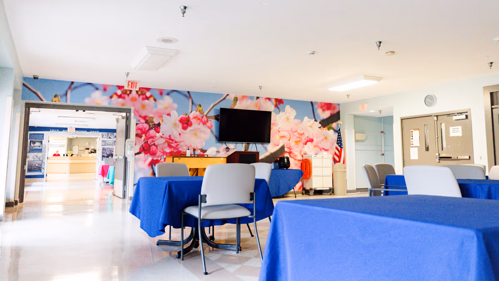 Interior view of a common area with tables covered in blue tablecloths and gray chairs. A large wall mural of pink cherry blossoms is visible behind a mounted flat-screen TV. The room has light-colored walls and floors, with an American flag in the corner and double doors leading to another area.