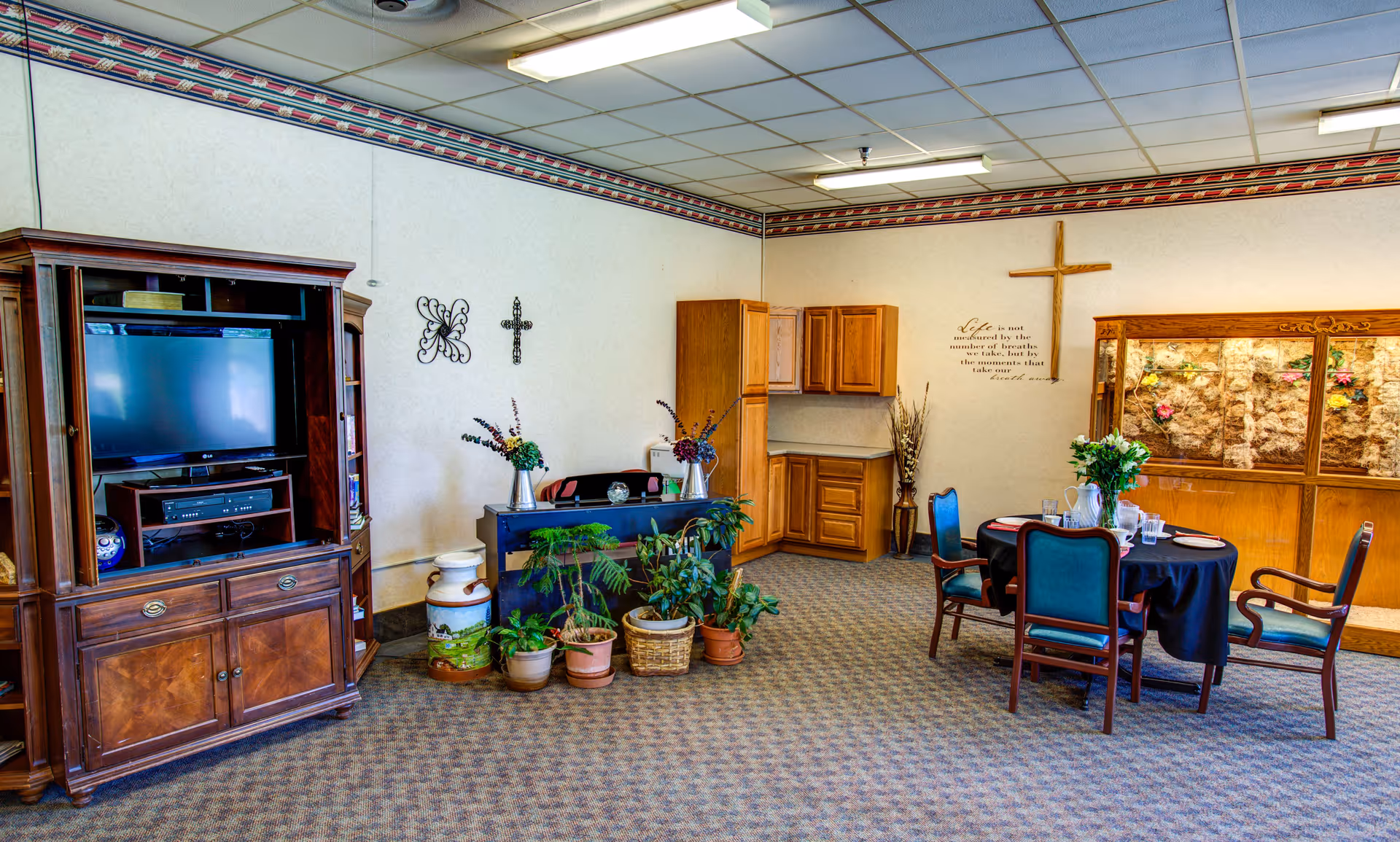 Community common room with a TV cabinet, potted plants, a small dining table with chairs, and religious wall decor.