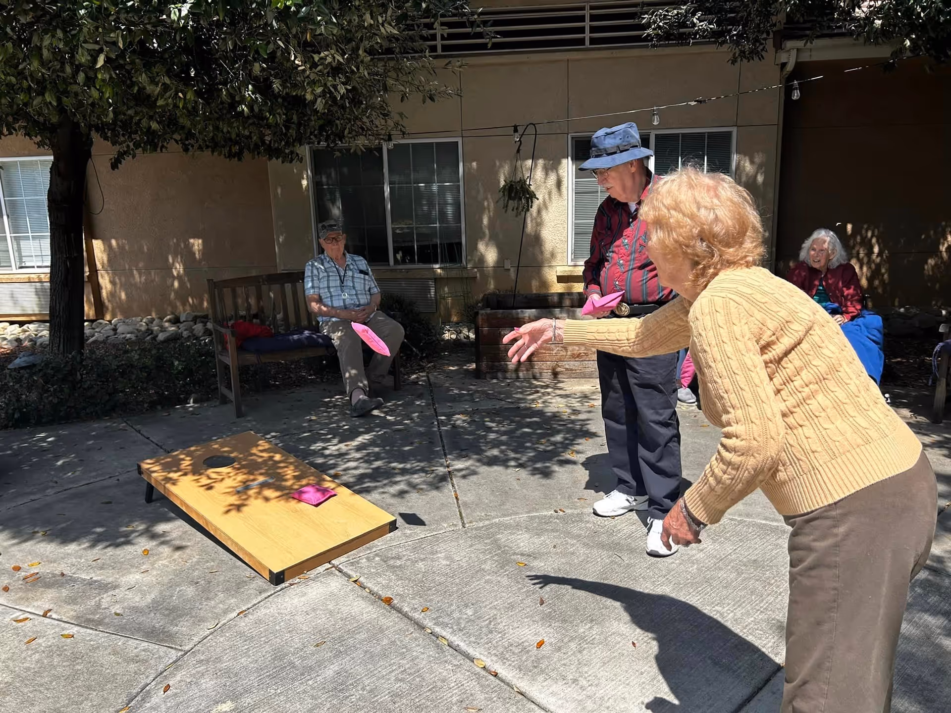 Four elderly people outdoors at Wildwood Canyon Villa Assisted Living and Memory Care playing a game of cornhole on a sunny day. One woman in a yellow sweater is throwing a pink bean bag towards the wooden cornhole board, while two men and another woman watch and participate. They are seated or standing on a concrete patio area with trees and a building in the background.