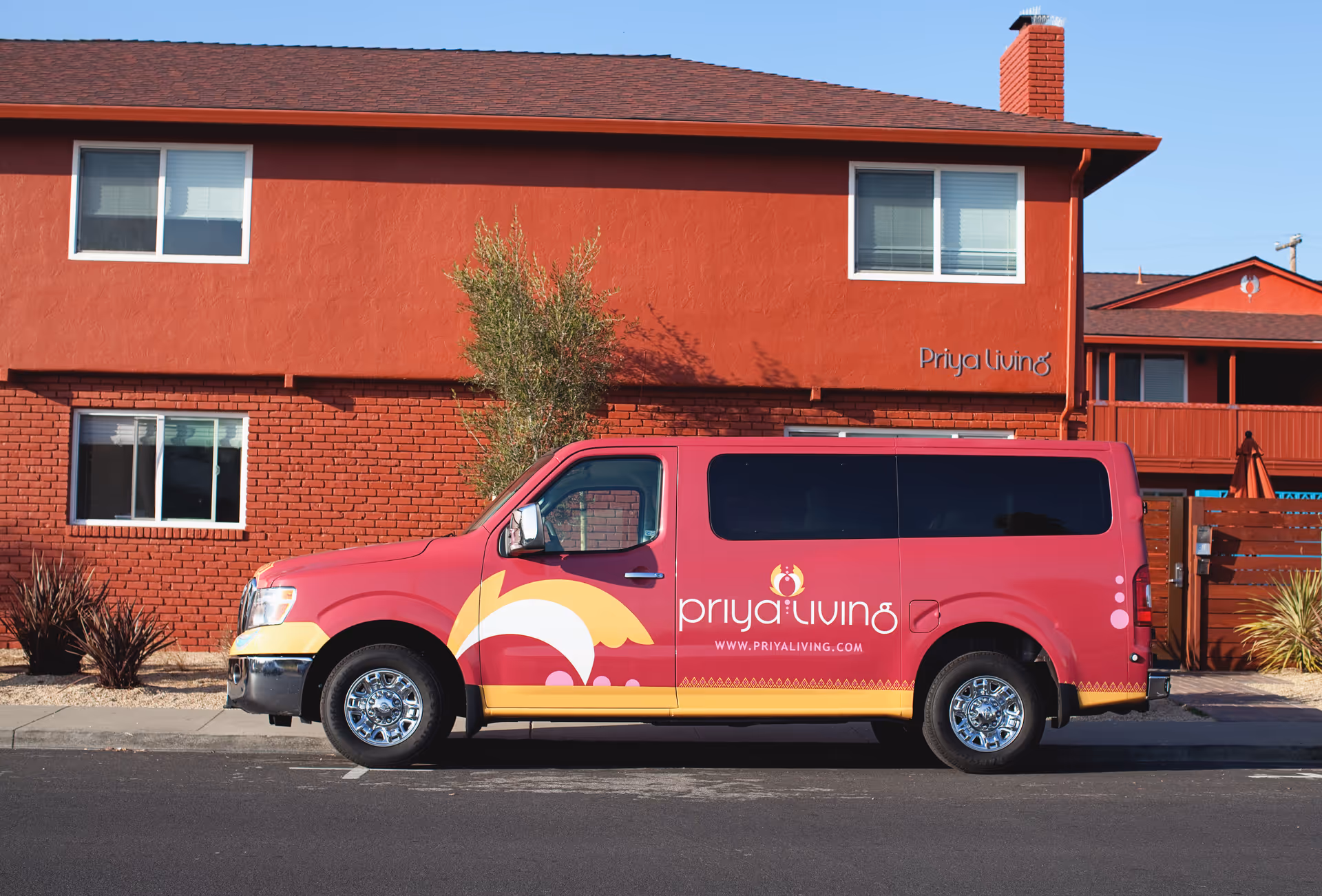 A red and yellow van with the logo and text 'priya living' and the website 'www.priyaliving.com' is parked in front of a two-story red brick and stucco building with the sign 'Priya Living' on the wall. The building has several windows and some landscaping with small plants and a tree.