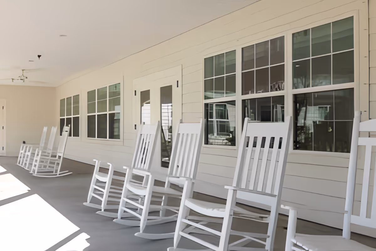 A covered porch area with several white wooden rocking chairs lined up along the wall of a building with multiple windows and a door.