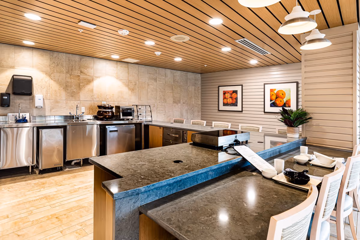 Modern kitchen area with stainless steel appliances, a large marble countertop island with seating, wooden ceiling with recessed lighting, and framed fruit artwork on the wall.