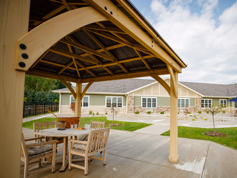 Outdoor patio area with a wooden gazebo covering a round table and four cushioned wooden chairs. The table has a tablecloth, a pitcher, glasses, and a plate of food. In the background, there is a single-story building with green and beige siding, stone accents, and multiple windows. The area is surrounded by grass, small trees, and paved walkways under a partly cloudy sky.