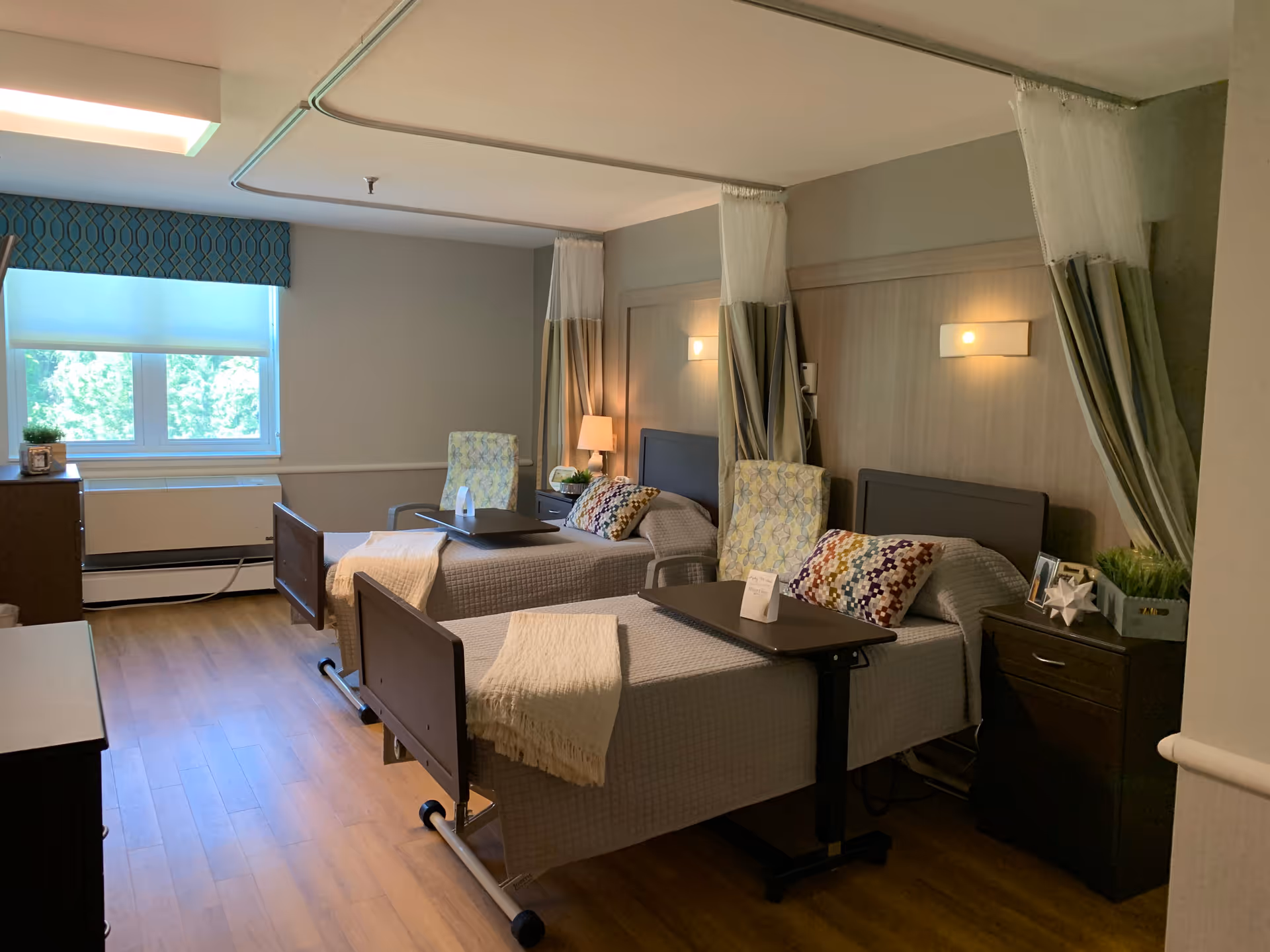 A shared patient room in a healthcare facility with two hospital beds, each with a colorful pillow and a beige blanket. Each bed has a small overbed table and a floral-patterned chair beside it. There are privacy curtains hanging from the ceiling around each bed. The room has wooden flooring, a window with a blue valance, and soft wall lighting above each bed. A nightstand with a lamp and decorative items is placed between the beds.