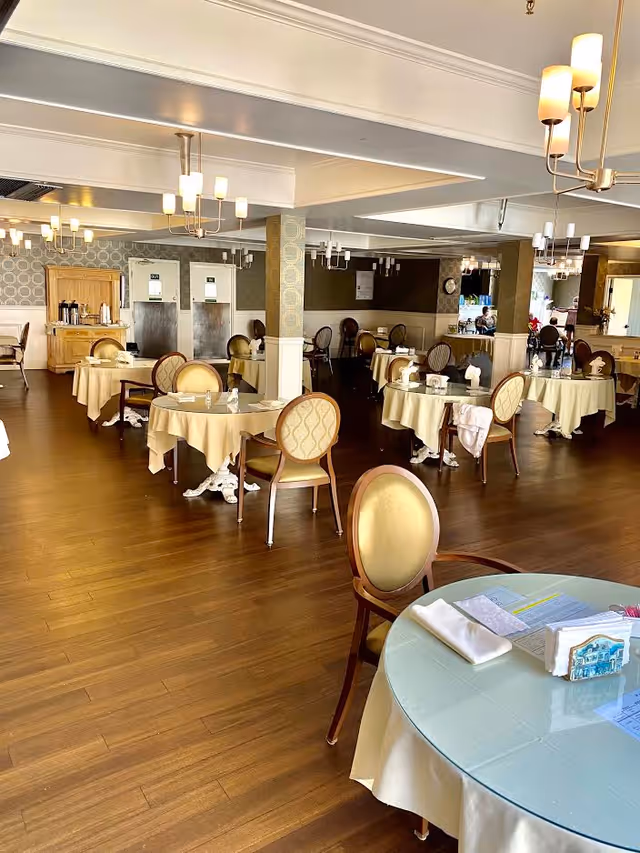 A spacious dining room with round tables covered in beige tablecloths, each set with napkins and glassware. The room features wooden flooring, elegant chairs with cushioned seats and backs, and multiple modern chandeliers hanging from the ceiling. The walls have decorative wallpaper and mirrors, and there is a wooden cabinet with coffee dispensers in the background.